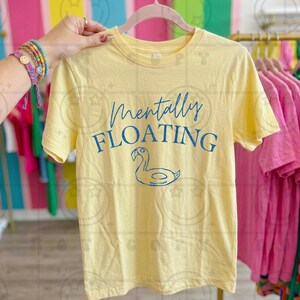 Peut inclure: T-shirt jaune clair avec l'inscription bleue "Mentally Floating" et un dessin de canard bleu. Le t-shirt est suspendu à un cintre rose.