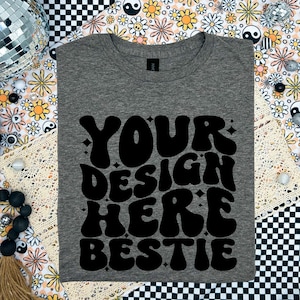 May include: A gray t-shirt with the text "YOUR DESIGN HERE BESTIE" in black, retro-style lettering.