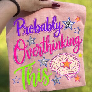 Puede incluir: Camiseta rosa claro con la frase "Probably Overthinking This" en morado, rosa y verde. El diseño incluye estrellas y un gráfico de cerebro con una estrella naranja en el centro.