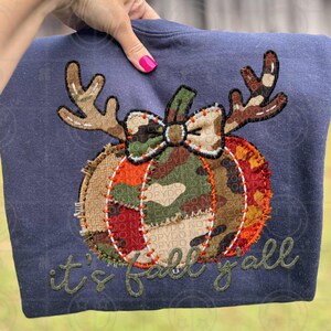 Peut inclure: Sweat-shirt bleu marine avec une applique de citrouille avec des motifs camouflage et jute, des bois et un nœud. L'expression "it's fall y'all" est brodée en vert.