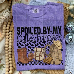 May include: A purple Comfort Colors t-shirt with the text "SPOILED BY MY Cattle Rancher" in black. The graphic includes cowboy boots, a hat, a jacket, and other ranching elements.