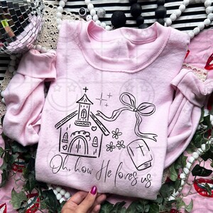 May include: A light pink sweatshirt with a crew neck and long sleeves. The front features a black line drawing of a church, a bow, flowers, and a book with a cross. The text "Oh, how He loves us" is below the drawing.
