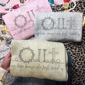 May include: Three sweatshirts in pink, grey, and cream, each with a black line drawing of a crown of thorns, nails, and a cross. The text "Out" and "we love because He first loved us" are printed on the sweatshirts.