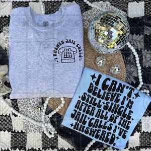 May include: Two t-shirts with humorous text. One is light gray with a telephone graphic and the text "I ANSWER JAIL CALLS." The other is light blue with the text "I CAN'T BELIEVE I'M STILL SINGLE AFTER ALL OF THE JAIL CALLS I'VE ANSWERED."