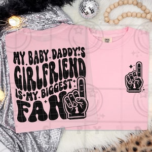 May include: Two light pink t-shirts. One shirt has the text "MY BABY DADDY'S GIRLFRIEND IS MY BIGGEST FAN" with a black and white foam finger graphic. The other shirt has a foam finger graphic with the number 1.