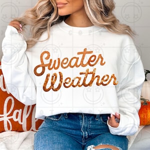 May include: A white long-sleeve sweatshirt featuring the text Sweater Weather printed in a metallic copper-coloured sequin-style graphic across the chest. The garment has a crew neckline and long sleeves with ribbed cuffs, paired with blue denim jeans.