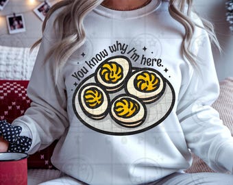 May include: White crew neck sweatshirt with a graphic of a plate of devilled eggs. The text reads "You know why I'm here." The design features four eggs with yellow yolks and a black outline.