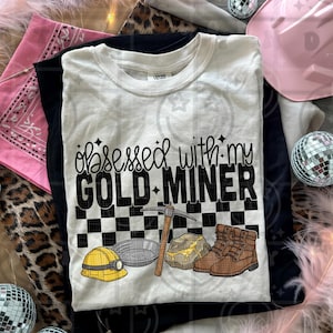 May include: White t-shirt with the text "Obsessed with my Gold Miner" in black script and block letters. The design includes a checkerboard pattern, pickaxe, gold pan, gold nugget, boots, and a yellow hard hat.