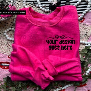 Puede incluir: Una sudadera rosa brillante con el texto "your design goes here" en negro. La prenda de cuello redondo está doblada, mostrando el área de diseño. El fondo incluye elementos decorativos como cuentas y vegetación.