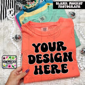 May include: A bright orange t-shirt with the text "YOUR DESIGN HERE" in black, retro-style font. The shirt is folded and laid on a white fuzzy surface.