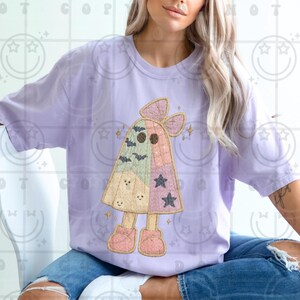May include: A light purple t-shirt featuring a whimsical ghost design. The ghost is made of patchwork fabric in pastel colors, with a bow, stars, and bats. The ghost is wearing pink boots. The shirt is a comfortable, casual style.