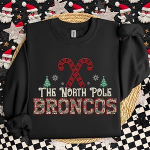Può includere: Felpa nera con un design festivo. Presenta bastoncini di zucchero rossi e bianchi incrociati, fiocchi di neve e alberi di Natale. Il testo "THE NORTH POLE BRONCOS" è visualizzato in un carattere decorativo.