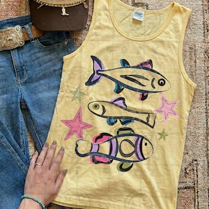 May include: Yellow tank top with a fish and star design. The sleeveless top features three colorful fish illustrations and several star shapes. The top is paired with blue jeans, a brown belt, and a brown purse.