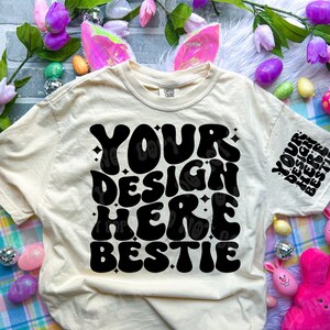 May include: A beige t-shirt with a black graphic that says "Your Design Here  Bestie" in a retro font. The shirt is surrounded by Easter decorations, including pink bunny rabbits, purple flowers, and colorful Easter eggs.