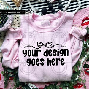 May include: A light pink sweatshirt with the text "your design goes here" in a black, bold font. The sweatshirt has a crew neck and long sleeves. The background includes decorative elements like beads and a disco ball.