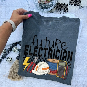 May include: Dark gray t-shirt with the words "future ELECTRICIAN" in black script and block letters. The shirt features a graphic of a yellow lightning bolt, pliers, a hard hat, a spool of wire, and a multimeter.