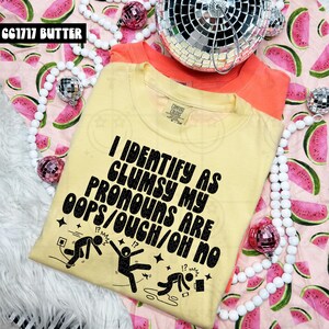 May include: A light yellow t-shirt with the text "I IDENTIFY AS CLUMSY MY PRONOUNS ARE OOPS/OUCH/OH NO" in black. The shirt has a crew neck and short sleeves. An orange t-shirt is behind the yellow shirt. The background has a watermelon pattern.