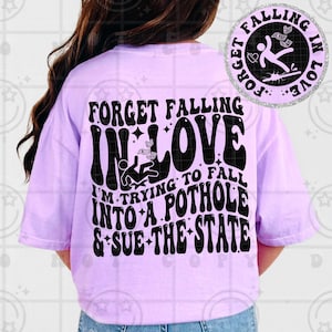 May include: A light purple t-shirt with the text "FORGET FALLING IN LOVE" and "I'M TRYING TO FALL INTO A POTHOLE & SUE THE STATE" in black. A circular graphic with the words "FORGET FALLING IN LOVE" surrounds a stick figure.