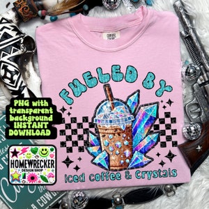 May include: Pink t-shirt with a graphic design of a cup of iced coffee and crystals. The text "Fueled by Iced coffee & Crystals" is printed on the shirt.