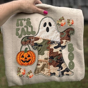 May include: Beige sweatshirt with a ghost applique holding an orange pumpkin. The ghost is made of camouflage fabric and white fabric. The words "IT'S FALL" and "BOO" are embroidered in green.