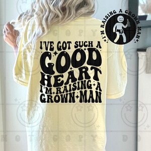 May include: A light yellow t-shirt with the text "I'VE GOT SUCH A GOOD HEART I'M RAISING A GROWN MAN" in black. A circular graphic on the upper right shows a baby holding a bottle. The shirt is a casual style.
