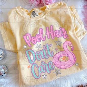 May include: A light yellow t-shirt with the phrase "Pool Hair Don't Care" in pink and teal lettering, along with a pink flamingo floatie and star graphics. The shirt is a Comfort Colors brand.
