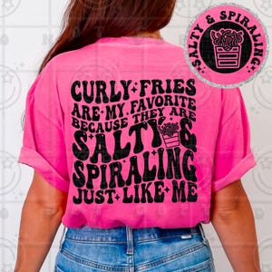 Può includere: Una maglietta rosa acceso con la scritta "Curly Fries Are My Favorite Because They Are Salty & Spiraling Just Like Me" in nero, in stile retrò. Sul retro, una grafica circolare recita "Salty & Spiraling" con un'illustrazione di patatine fritte.