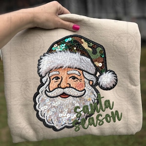 May include: A beige sweatshirt featuring an embroidered Santa Claus face with a sequined beard and a camouflage hat. The words "Santa Season" are embroidered in green script below the image.