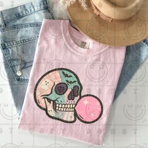 May include: A light pink Comfort Colors t-shirt with a skull graphic and bubblegum bubble. The skull design features pastel pink, blue, and purple sections, bats, and ghosts. A tan hat and blue jeans are in the background.