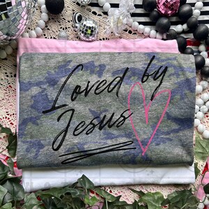 May include: Three folded t-shirts in pink, camouflage, and white. The camouflage shirt features the text "Loved by Jesus" in black script with a pink heart. The shirts are stacked on a decorative surface with beads and disco balls.