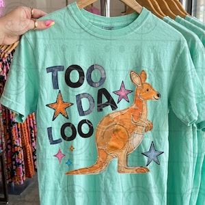 May include: A light teal t-shirt featuring the text "TOO DA LOO" with star accents and a kangaroo applique. The kangaroo is orange and tan, with a patchwork design. The shirt is short-sleeved and has a crew neck.