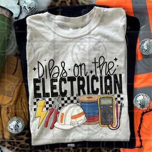 May include: Cream-colored t-shirt with the text "Dibs on the Electrician" in black. The design includes illustrations of a lightning bolt, pliers, a hard hat, a spool of wire, and a multimeter. The shirt is folded on top of a black shirt.