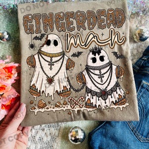 May include: A tan t-shirt featuring a graphic of two gingerbread ghosts with icing, wearing chains and crosses. The text reads GINGERDEAD man in a brown, decorative font. The design includes small bats, a spider, and candy canes.
