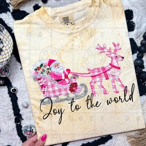 May include: Cream-colored t-shirt featuring a festive design with Santa Claus in a pink plaid sleigh pulled by a pink plaid reindeer. The text "Joy to the world" is written in cursive. The shirt has a Comfort Colors tag.