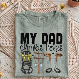 Op de afbeelding: Een lichtgroen t-shirt met de tekst "MY DAD climbs Poles" in het zwart. Het shirt heeft illustraties van een klimharnas, een elektriciteitspaal en klimsporen. Het shirt is gemaakt van een zacht materiaal.