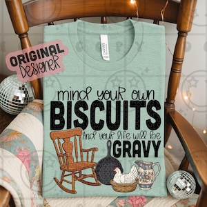 Può includere: Maglietta verde chiaro con la scritta "mind your own BISCUITS and your life will be GRAVY". Il design include una sedia a dondolo, una padella, una gallina in un cestino e una brocca. La maglietta è in materiale morbido.
