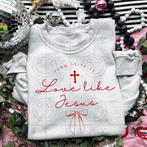May include: A light gray sweatshirt with the text "John 13:34-35" above a red cross and the words "Love like Jesus" in a cursive font. A red bow is below the text. The sweatshirt has a crew neck and long sleeves.
