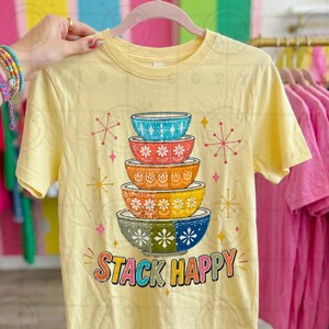 May include: Yellow t-shirt featuring a stack of colorful, patterned bowls with the text "STACK HAPPY" in a retro font. The bowls have floral and geometric designs in shades of blue, orange, yellow, and green. Pink and yellow starbursts and stars are scattered around the bowls.