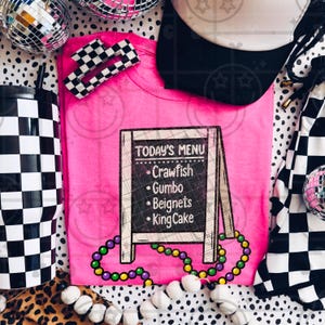 Könnte beinhalten: Ein leuchtend pinkfarbenes T-Shirt mit einer Tafel im Kreidetafel-Stil, auf der "Crawfish", "Gumbo", "Beignets" und "King Cake" stehen. Das Shirt ist mit einer Mardi Gras-Perlenkette und einer schwarz-weiß karierten Haarspange verziert.