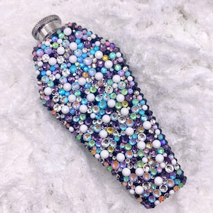 May include: A black, coffin-shaped flask covered in iridescent rhinestones. The rhinestones are in various colors, including white, purple, blue, and green.