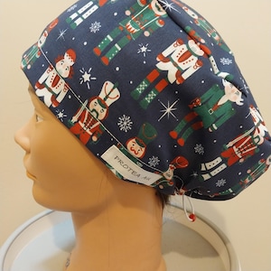 May include: A navy blue scrub cap with a repeating pattern of red, green, and white toy soldiers and snowflakes. The cap has a white tag with the text "PROTEA AU". The cap is designed for use in a medical environment.