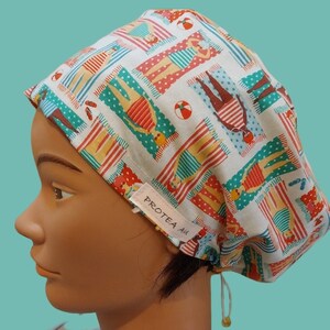 May include: A white scrub cap featuring a repeating pattern of people lounging on beach towels with beach balls. The fabric has a small white tag on the side with the text PROTEA AU printed in black. The cap includes a drawstring with a toggle.