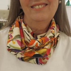 May include: A lightweight infinity scarf featuring a cream-coloured base with a vibrant floral pattern in shades of orange, red, yellow, purple, and green. The fabric is draped in multiple folds around the neck, showcasing a smooth, soft texture.