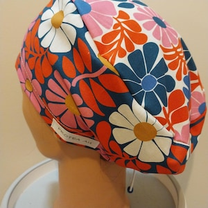 May include: A patterned scrub cap with a floral design. The cap displays large flowers in white, pink, and blue, with orange leaves on a dark teal backdrop. The tag reads "PROTEA AU".