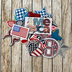 May include: A collection of patriotic patches and embellishments in red, white, and blue. Includes an American flag, stars, hearts, and the word "AMERICA" in a decorative font. The items are arranged on a wooden background.