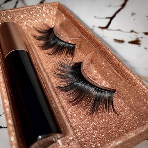 Könnte beinhalten: Ein Set aus schwarzen falschen Wimpern und einer schwarzen Eyeliner-Tube mit kupferfarbener Kappe, präsentiert in einer roségoldenen Glitzerbox. Die Wimpern sind ordentlich angeordnet und zeigen ihr dunkles, voluminöses Aussehen. Kosmetikverpackung.