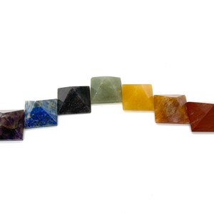 May include: Set of seven chakra crystal pyramids in various colors, including purple, blue, green, yellow, orange, red, and brown. The pyramids are arranged in a semi-circle.