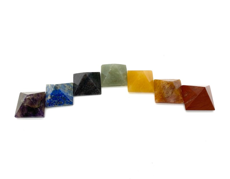 May include: Set of seven chakra crystal pyramids in various colors, including purple, blue, green, yellow, orange, red, and brown. The pyramids are arranged in a semi-circle.