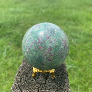 Peut inclure: Une sphère de zoïsite rubis verte et rose sur un support doré. La sphère est polie et a une surface lisse.