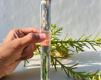 May include: A clear, faceted crystal wand with a pointed tip and a geometric design. The wand is held by a hand, with green foliage in the background. The crystal wand is approximately 7 inches in length.